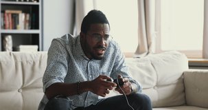 Happy emotional young adult african american guy gamer player holding gamepad controller playing feel excited winning video game sit alone on couch at home, console videogames hobby concept