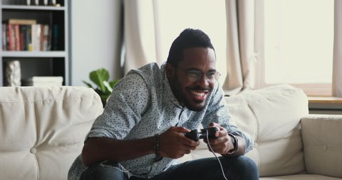 Happy emotional young adult african american guy gamer player holding gamepad controller playing feel excited winning video game sit alone on couch at home, console videogames hobby concept