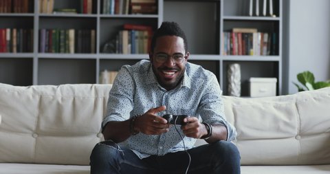 Funny excited young adult african mixed race man gamer winner holding controller playing videogame sitting on sofa feeling overjoyed celebrating victory winning in video game alone at home