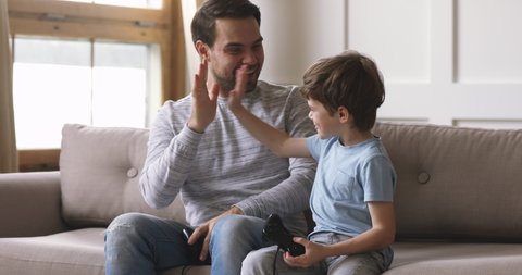 Overjoyed young adult dad and cute child son gamers winners playing winning video game, excited father having fun with kid boy give high five holding celebrating videogame victory at home