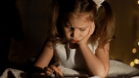 Baby girl lies in bed and plays on smartphone in a game in the dark. Close up face of the child is lit by a bright screen.