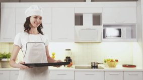 Attractive young woman and her little cute daughter in kitchen aprons and hats cooking biscuits together. Young lady holding plate with cookies to put them in oven