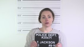 Mugshot of Female Criminal stood against a height chart. The Woman is dressed in an greay and holding an ID board  -4K Stock Video clip footage