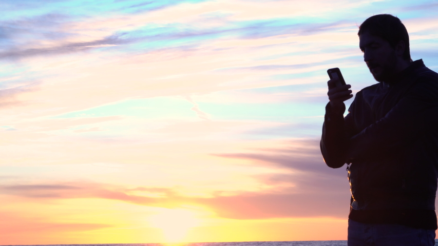 A man watches a photo on social networks at sunset by the sea.