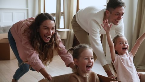 Excited happy young parents run push cardboard boxes with little cute children daughters ride inside, family homeowners playing having fun on moving day celebrate mortgage relocation removals concept