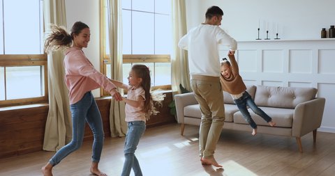 Happy active family young adult parents mum dad and cute little children daughters holding hands dancing jumping together in living room interior enjoying funny weekend activity in modern apartment