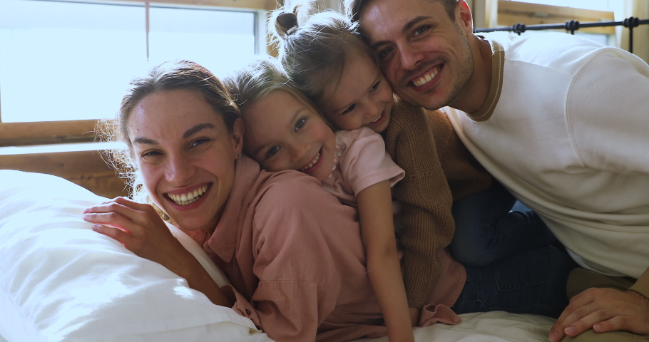 Happy funny beautiful family of four bonding laughing relaxing on comfortable bed looking at camera, cheerful young parents with cute little kids daughters lay in bedroom in morning, closeup portrait Royalty-Free Stock Footage #1044147385