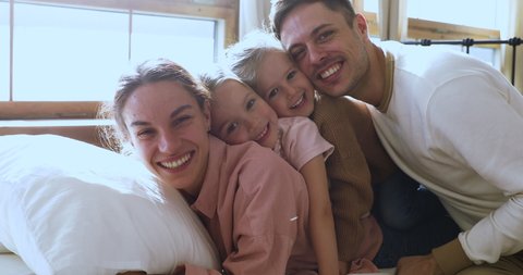 Happy funny beautiful family of four bonding laughing relaxing on comfortable bed looking at camera, cheerful young parents with cute little kids daughters lay in bedroom in morning, closeup portrait