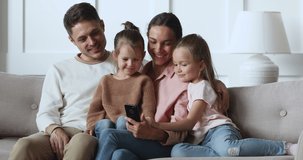 Smiling parents and small children talking using smart phone gadget relax together on sofa, happy family mom dad little daughters hold look at smartphone doing shopping in mobile app on couch at home