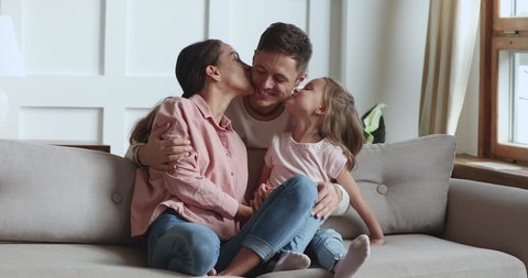 Happy young husband father hugging wife and cute little child daughter kiss dad on cheeks look at camera, beautiful family with small kid bonding laughing sit on sofa, fathers day concept, portrait