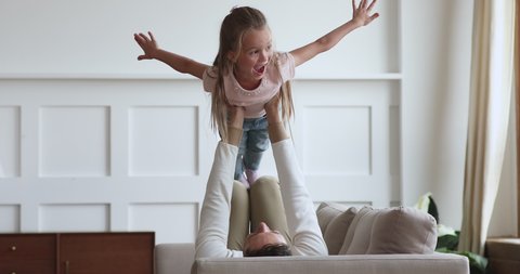 Cute excited child girl play plane game fly in fathers arms spend leisure time with parent at home, happy dad lifting little kid daughter up bonding together lounge on sofa enjoy lifestyle activity