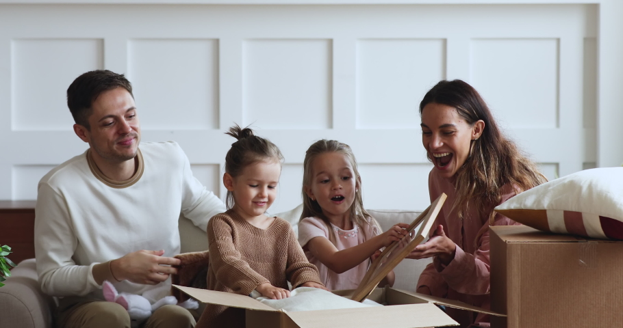 Excited family with two cute little kids daughters unpacking boxes sit on sofa in new apartment on moving day, young parents and small children customers open parcels buying home furnishings concept Royalty-Free Stock Footage #1044147448