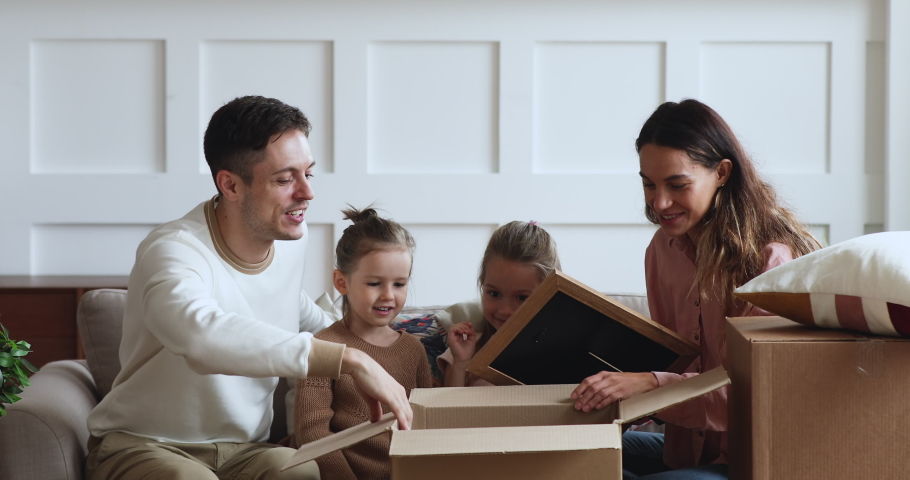 Excited family with two cute little kids daughters unpacking boxes sit on sofa in new apartment on moving day, young parents and small children customers open parcels buying home furnishings concept Royalty-Free Stock Footage #1044147448