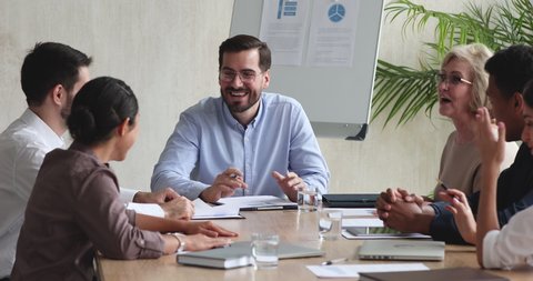 Happy male ceo boss manager telling funny joke having fun talking with multiethnic employees during group briefing, cheerful friendly diverse business team people laughing sitting at meeting table