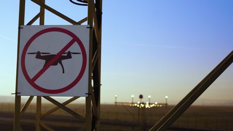 No drone zone sign on approach lighting system at runway. Airport airspace perimeter prohibition drones fly sign. Airport infrastructure and buildings in background, against blue sky.