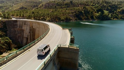 Side aerial drone shot of white modern camping van with bike transportation racks drive over bridge or dam road in blue waters.Free and inspired woman hangs out of window. Happy and carefree lifestyle