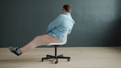 Young man with expressive face is spinning on office chair gesturing making funny grimaces sitting alone moving on dark background. People and fun concept.