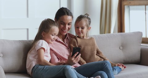 Happy young mum using social media app on smartphone recording video blog having fun with two cute small children daughters embracing bonding together looking at mobile phone relaxing on sofa at home