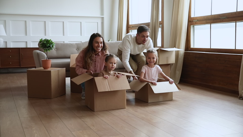 Funny active family playing on moving day, excited happy adult parents mom dad pushing cardboard boxes with cute little kids sit inside having fun packing relocate into new home concept, slow motion Royalty-Free Stock Footage #1044184111