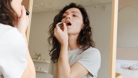 Young female holding lipstick in her hands and paints lips prepare getting ready in the morning. She looks at herself in the mirror and smiles. Woman doing makeup. Get ready for a date.
