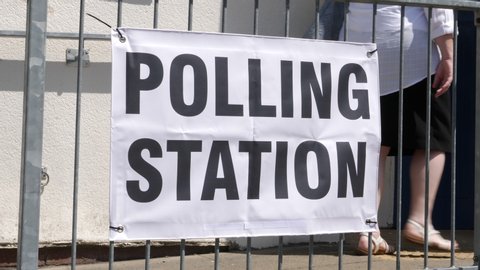 Polling Station Sign on railings - People walking past and going to vote in the election. Politics. Slow Motion