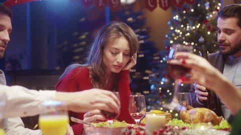 Lonely sad young woman sitting in frustration near Christmas tree gathering with happy friends at dinner party. Sad Christmas story. Loneliness concept.