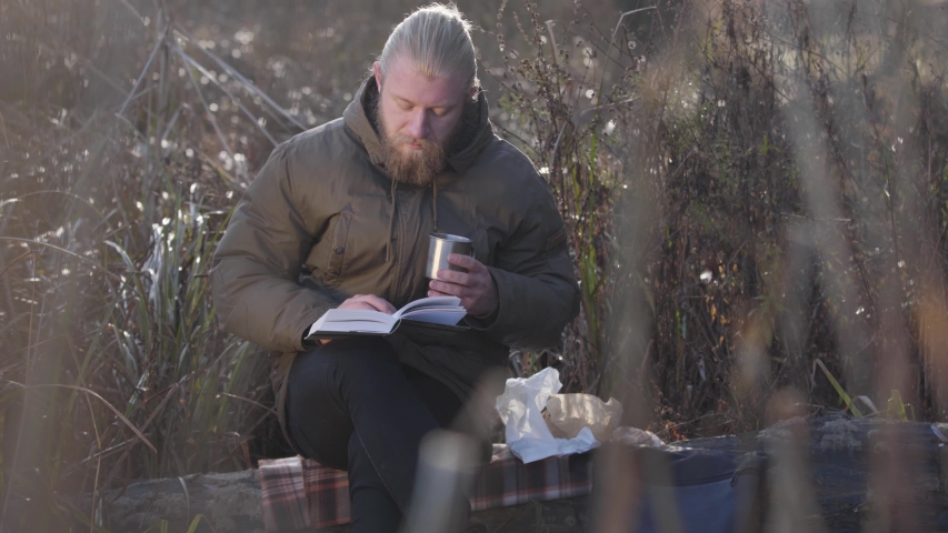 Thoughtful Caucasian man sitting on tree trunk, reading and drinking hot tea from steel mug. Lonely male tourist enjoying resting outdoors. Leisure, lifestyle, hobby.