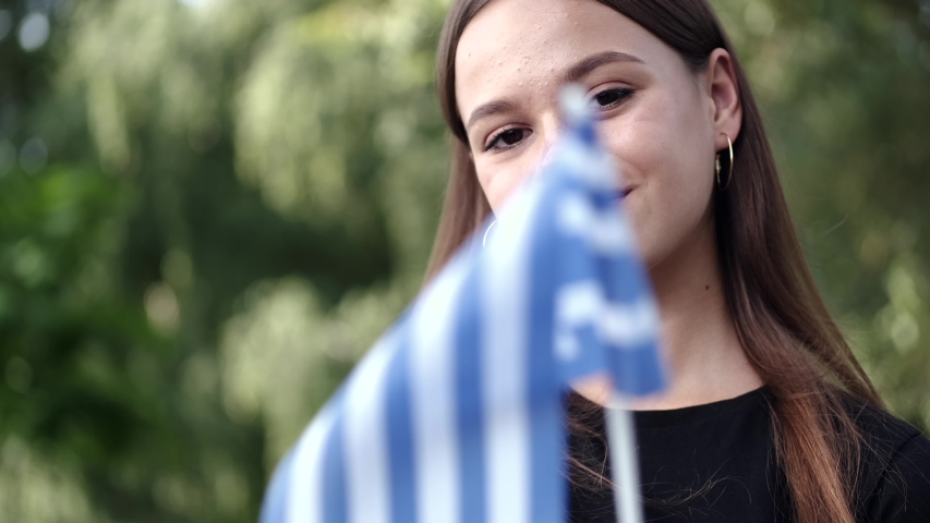 A young lady is showing the Greek national flag. The national symbol is waved on a stick. The girl is standing at the park.