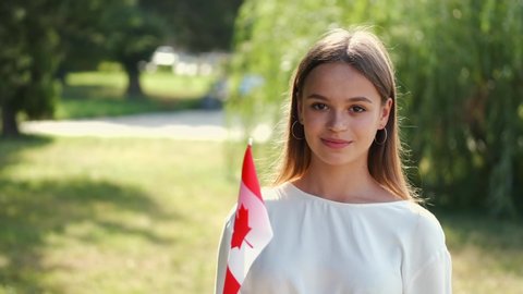 A teen is waving the flag of Canada. The girl is smiling and standing at the park.