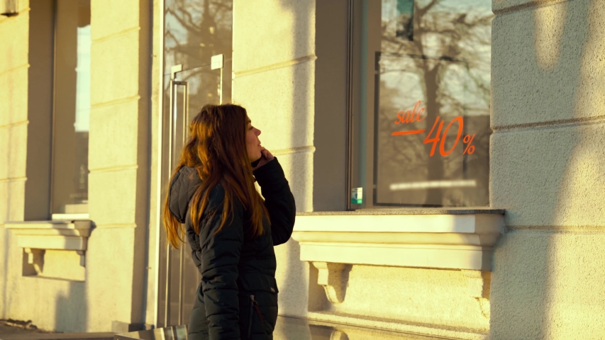 Young girl talking on phone and looking at shop window with sale sign
