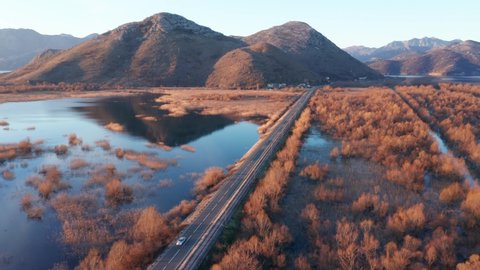 Winter sunrise panorama of Lake Skadar toward Vranjina island and hill. Flying over the main road connecting Podgorica and the Adriatic coast. Bare trees and blue sky reflection in the water.