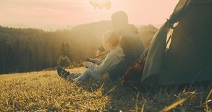 Happy traveler father and son sitting on grass and look through binoculars, bonding around mountain under sun light enjoying the leisure and freedom. 4K slow motion video
