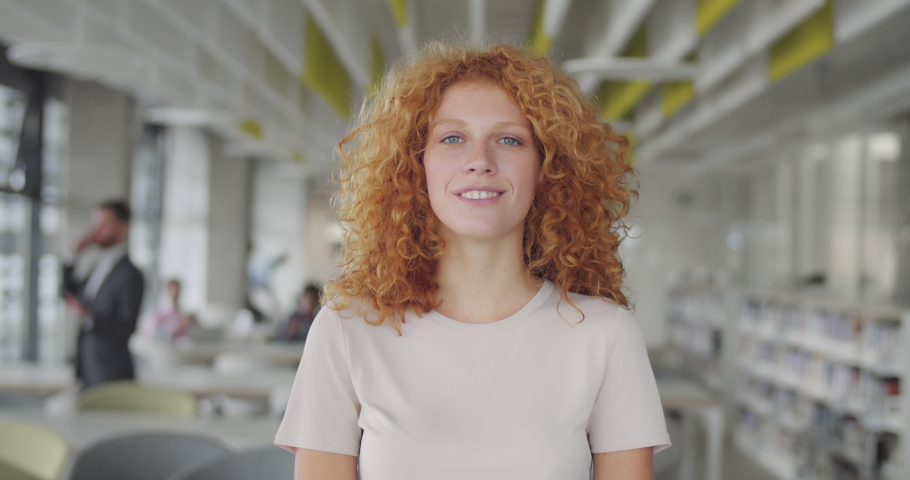 Front view of beautiful young female professional administrator looking to camera and smiling. Young girl office worker standing in busy modern office. Positive emotions. Royalty-Free Stock Footage #1044237706
