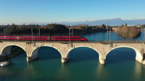 High-speed train moving on an arch bridge over the Mincio River in the city of Peschiera del Garda, Italy. In the background the Alps in the snow. The clear blue sky. Bridge reflection in water