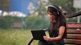 Young woman on bench with headphones and laptop listening to music. Outdoors