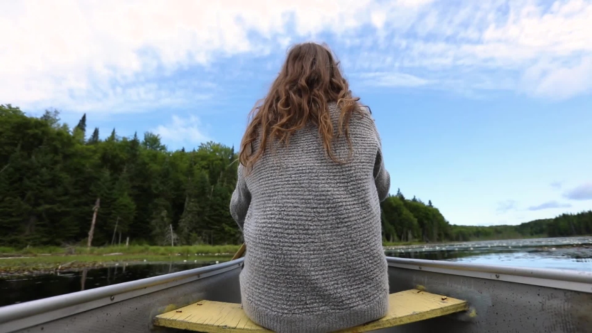 Scenic and mindful canoe voyage as a brunette woman is seen from the back, sitting on narrow boat using a wooden paddle to row on a large lake