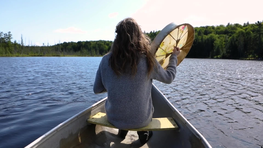 Slow motion footage onboard a narrow canoe over a scenic lake, as a woman sits up front playing a shamanic drum. Mindful retreat in Quebec, Canada Royalty-Free Stock Footage #1044244681