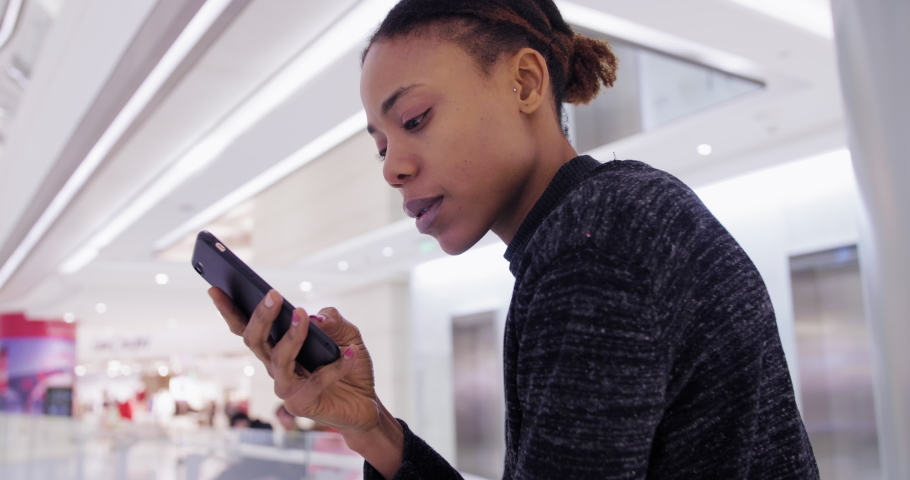 Side view of one happy young African woman browsing web on mobile phone in shopping center black girl using looking at smartphone in urban mall young people lifestyles with cell 4k clip