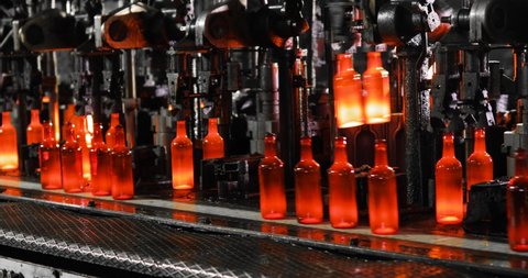 Automated process of forming bottle of molten glass. Molten glass mass enters mold and then into glass blower where bottle is formed. Production of glass bottles on manufacturing industrial factory.