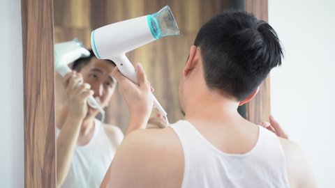 Back view of Asian guy in white shirt blowing dry hair using hairdryer in front of mirror