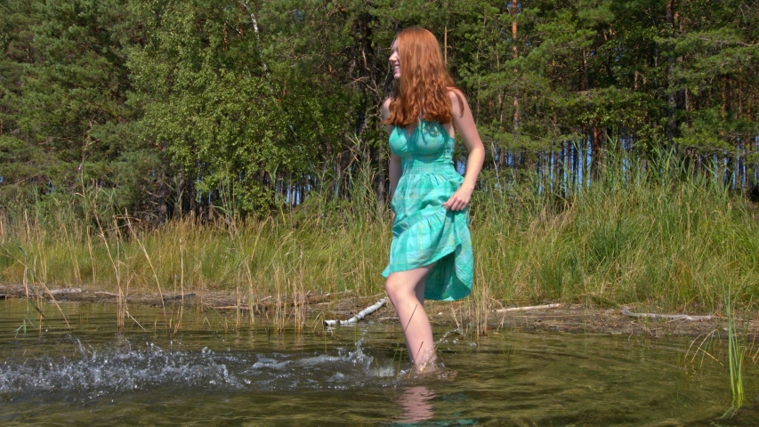 Happy Beautiful ginger hair woman is having fun while kicking the water of a calm lake, smiling and looking far away the camera with forest trees as background during the day
