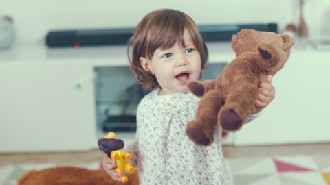 Portrait of cute little one year old girl laughing happily while playing and hugging with her mother at home with a teddy bear. Shot in 6k, cine lens. Instagram style.