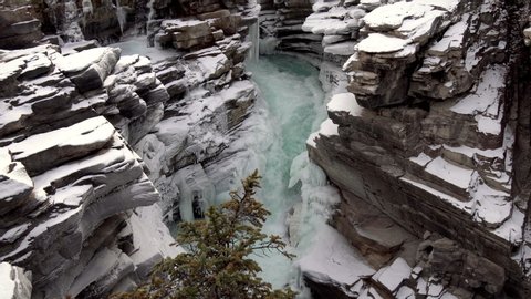 Nearly frozen waterfall with pine trees in the foreground and beautiful teal colored water, Jasper National Park, Alberta Canada, Aerial pan right shot