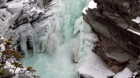 Raging water in a nearly frozen waterfall with pine trees in the foreground, Jasper National Park, Alberta Canada, Aerial pan right shot