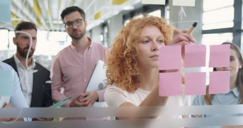Close up of female office worker using glass board and proposing her problem solving to workmates. Diverse coworkers having corporate meeting and standing near glass wall with sticky notes.