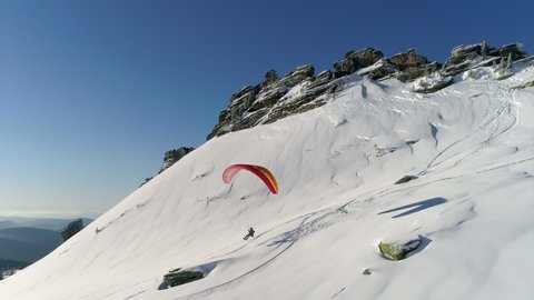 AERIAL: epic video of a paraglider flying in the mountains in winter and descending on the background of a snowboarder from a sheer cliff wall. snowboard and parachute in the mountains in winter