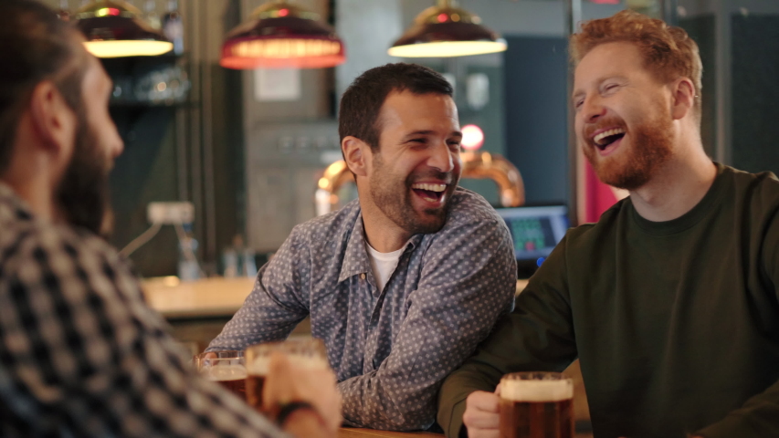 Smiling carefree friends enjoying drinking together in bar. Group of happy young men drinking cold draft beer, chatting and having good time at pub. Laughing men on night out drinking beer on counter. Royalty-Free Stock Footage #1044263386