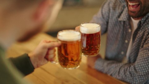 Closeup of two hands clinking beer stein glasses. Toasting with a pint of draft beers on counter at pub. Detail of happy men hands raising toast with cold beer mugs at bar.