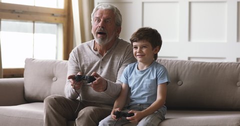 Emotional happy old senior grandpa and cute little kid grandson gamers team hold controllers play video game sit on sofa, two age generations family having fun enjoy videogame at home