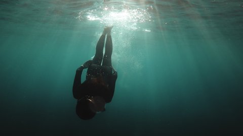 Redhead girl in a black neoprene suit splashes and swims underwater. Sun rays in clear emerald water. Spray and sun glare on the sea sand bottom. Swimming in open water. Beautiful underwater landscape